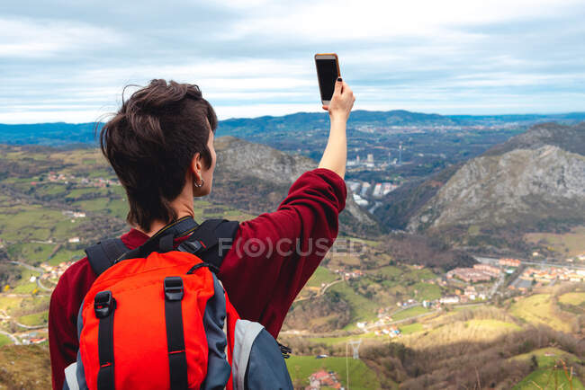 Back view of unrecognizable woman with backpack standing with arm raised and taking shot with smartphone of wonderful scenery with small villages and town in valley against foggy ridges at horizon under cloudy sky in Asturias — Stock Photo