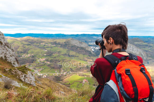 Vue latérale de la femme avec sac à dos debout sur la clairière et prise de vue avec caméra de magnifique vallée contre les crêtes brumeuses à l'horizon sous le ciel avec des nuages pelucheux en Espagne — Photo de stock