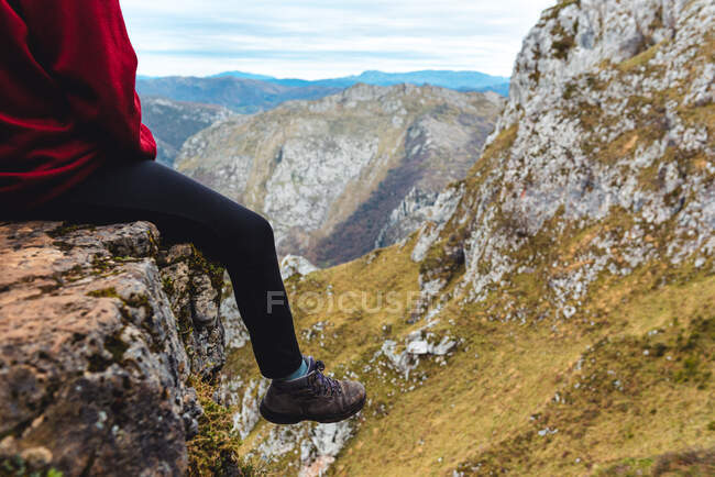 Vista lateral de las piernas turista sentado en el borde del acantilado disfrutando de la libertad y admirando increíbles paisajes de campo situado en el valle al pie de la montaña contra las colinas boscosas brumosas y llanura bajo el cielo con nubes grises exuberantes en España - foto de stock