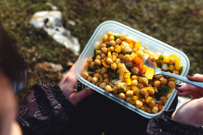 From above crop woman in warm jacket holding plastic container of chickpea with veggies having lunch on meadow while hiking in Spain — Stock Photo