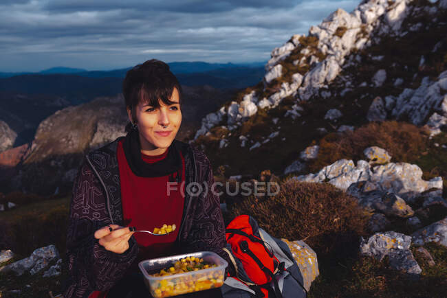 Woman in warm jacket holding plastic container of chickpea with veggies having lunch on meadow while hiking in Spain — Stock Photo