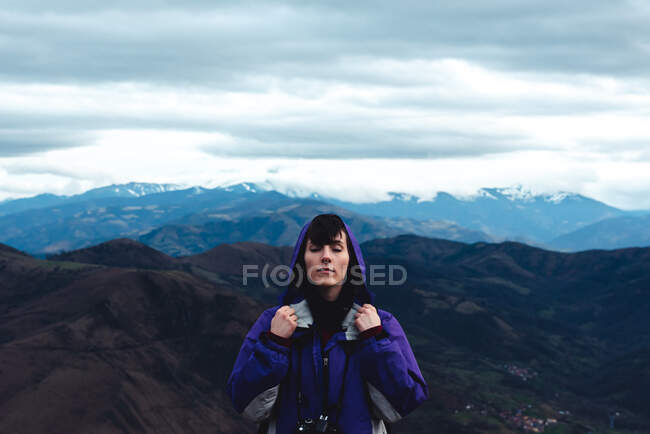 Woman tourist in violet jacket with backpack with close eyes in picturesque mountain ridges under cloudy sky while standing in Monsacro — Stock Photo