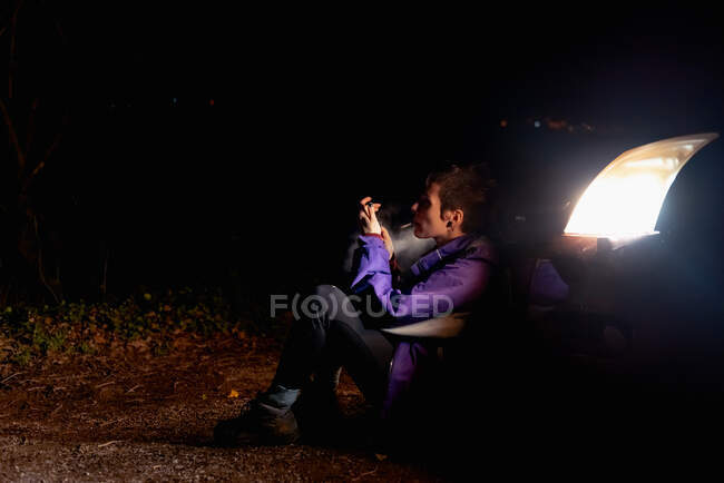 Провокационная уверенная женщина в фиолетовой и яркой оранжевой куртке, зажигая сигарету, сидя в одиночестве, опираясь на автомобиль с яркими фарами в темную ночь в Испании — стоковое фото