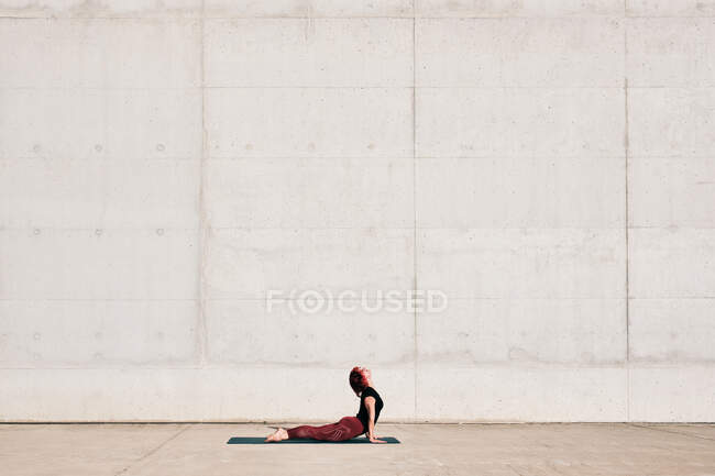Vista laterale di atleta femminile alla moda in abbigliamento sportivo che fa esercizio di yoga cobra bambino sul tappeto sportivo mentre si allena da solo sulla strada — Foto stock