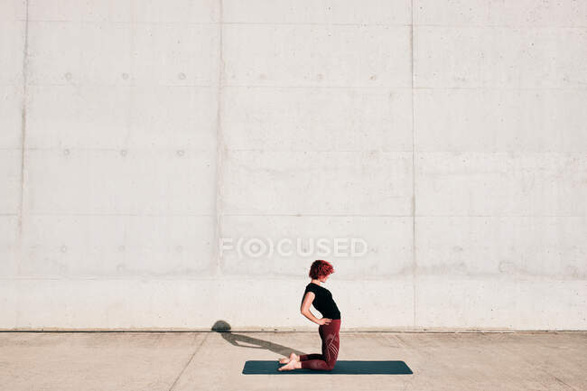 Donna alla moda in abbigliamento sportivo facendo yoga in posa cammello sul tappeto sportivo da solo sulla strada contro il muro di cemento nella giornata di sole — Foto stock