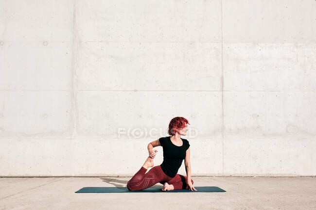 Vista lateral de atleta femenina en ropa deportiva sentada en badha hamsasana en alfombra deportiva y mirando hacia otro lado mientras practica yoga - foto de stock