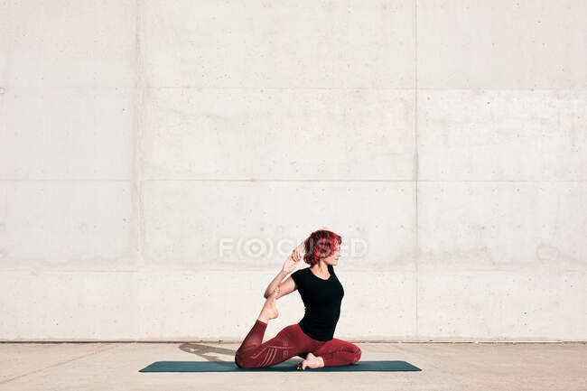 Vista laterale di atleta donna in abbigliamento sportivo seduto in badha hamsasana sul tappeto sportivo e guardando lontano durante la pratica dello yoga — Foto stock