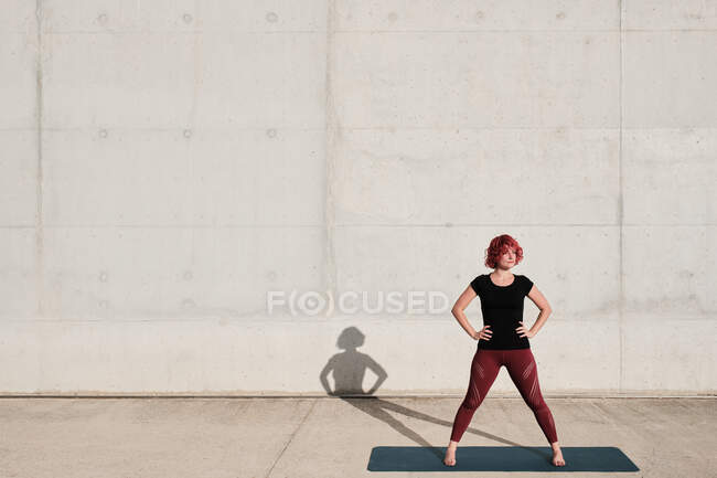Подходит вдумчивая женщина с вьющимися рыжими волосами в спортивной одежде созерцая, стоя на коврике для йоги против бетонной стены — стоковое фото