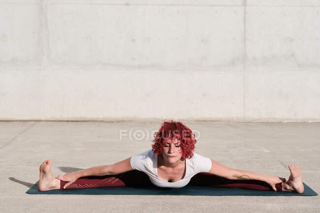 Desde arriba de mujer descalza con los ojos cerrados en ropa deportiva haciendo yoga en amplio ángulo sentado hacia adelante pose de flexión en la estera - foto de stock