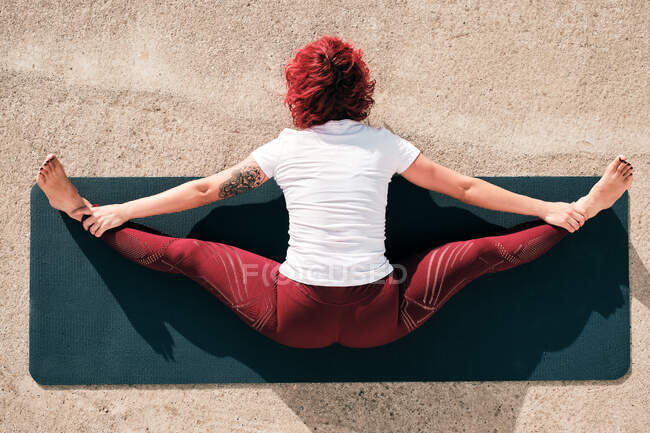 Сверху вид сзади на анонимную босоногую женщину в спортивной одежде, занимающуюся йогой под широким углом сидящую вперед позу изгиба на коврике — стоковое фото