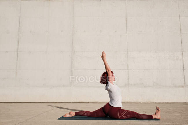 Vue latérale de la femme pieds nus avec les yeux fermés en vêtements de sport faisant du yoga en pose de singe sur la formation de tapis seul dans la rue — Photo de stock