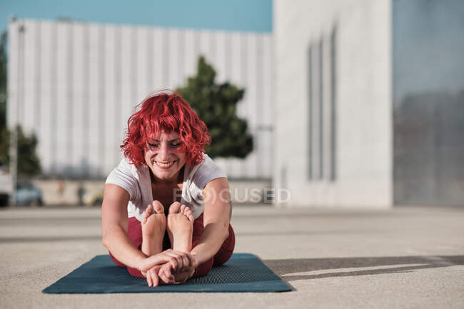 Deportista femenina descalza flexible con el pelo rizado rojo en ropa deportiva sentada en paschimottanasana y sonriendo mientras practica yoga en la calle - foto de stock