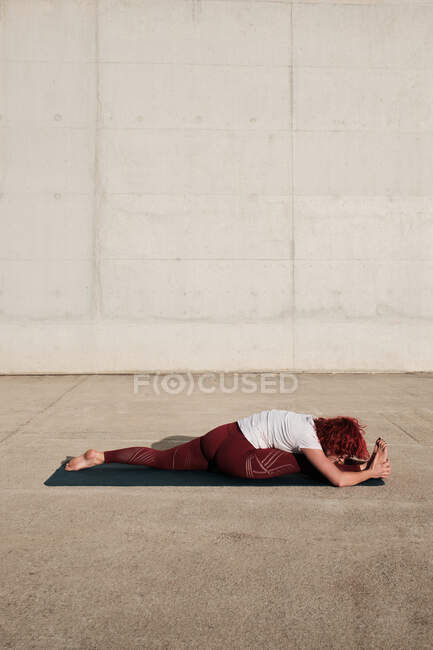 Vista lateral de mujer descalza anónima en ropa deportiva haciendo yoga en mono pose hacia adelante pose de flexión en la estera - foto de stock