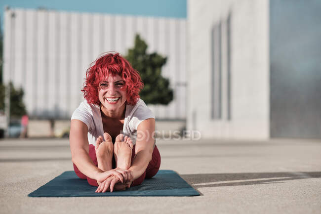 Гибкая босоногая спортсменка с рыжими вьющимися волосами в спортивной одежде, сидящая в пасхимоттанасане и улыбающаяся во время занятий йогой на улице — стоковое фото