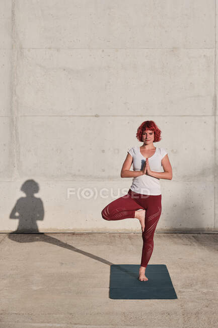 Уверенная босиком спортсменка в спортивной одежде медитирует, стоя в позе врикшасаны с закрытыми к груди руками в намасте — стоковое фото