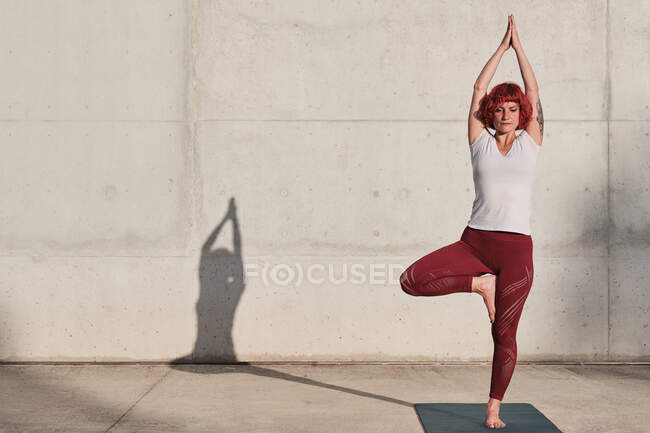 Confident barefooted female athlete in sportswear meditating while standing in vrikshasana position with arms raised and namaste — Stock Photo