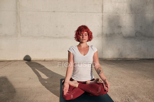 Mulher com cabelo vermelho e tatuagem em camisa branca e calças bordô sentado em padmasana com Gyan Mudra e meditando com olhos fechados — Fotografia de Stock