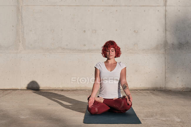 Mujer con pelo rojo y tatuaje en camisa blanca y pantalones borgoña sentada en padmasana con Gyan Mudra y meditando con los ojos cerrados - foto de stock