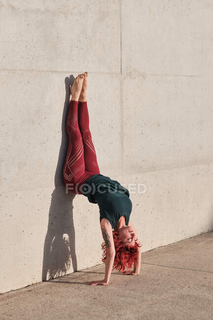 Vista lateral da fêmea descalça magro com cabelo vermelho em sportswear de cabeça para baixo em pose de cão voltado para baixo inclinado na parede de concreto — Fotografia de Stock