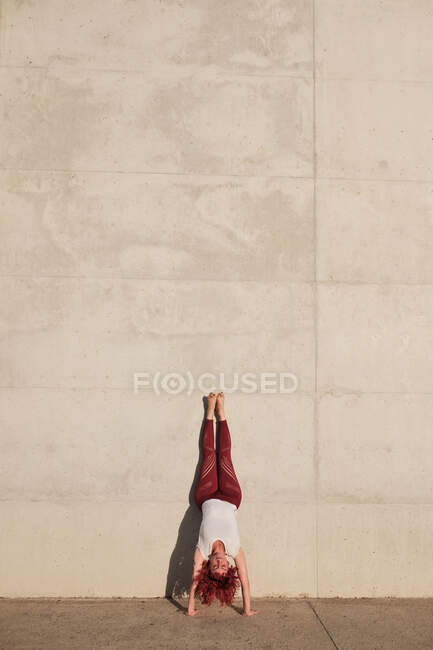 Femme mince pieds nus avec des cheveux roux dans les vêtements de sport debout à l'envers dans la pose de chien orientée vers le bas appuyé sur un mur en béton — Photo de stock