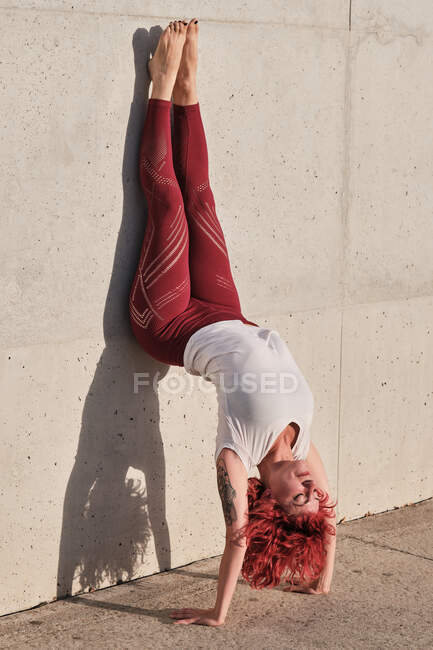 Vista lateral da fêmea descalça magro com cabelo vermelho em sportswear de cabeça para baixo em pose de cão voltado para baixo inclinado na parede de concreto — Fotografia de Stock