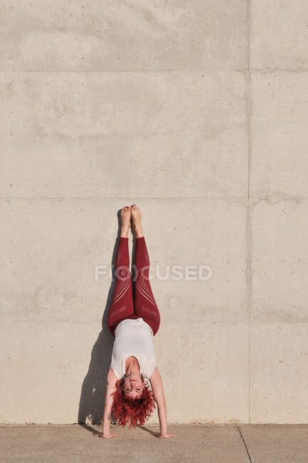 Тонкая босоногая самка с рыжими волосами в спортивной одежде, стоящая вверх ногами в позе собаки вниз головой, опираясь на бетонную стену — стоковое фото