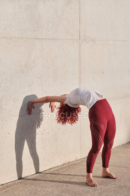 Slim hembra descalza con el pelo rojo en ropa deportiva de pie boca abajo en la postura hacia abajo perro apoyado en la pared de hormigón - foto de stock