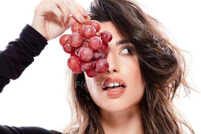 Gorgeous black haired lady in trendy clothing holding branch of red grapes in front of half face and looking away making funny faces in a isolated white background — Stock Photo