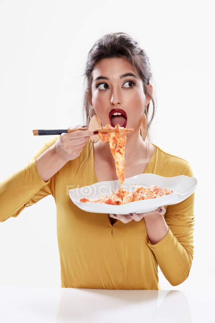 Funny young female with red lipstick looking away with opened mouth while tasting pasta with chopsticks isolated on white background — Stock Photo