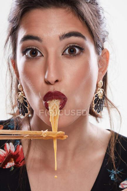 Femme à la mode avec un maquillage élégant en regardant la caméra avec des nouilles dans la bouche et en utilisant des baguettes isolées sur fond blanc — Photo de stock