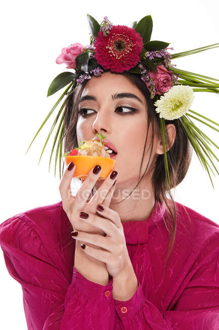 Mujer hermosa joven con corona de flores de colores en la cabeza mirando hacia otro lado mientras sostiene el plato en ralladura naranja aislado sobre fondo blanco - foto de stock