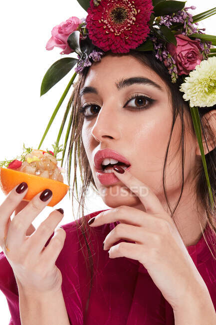 Молодая красивая женщина с венком красочных цветов на голове и пальцем на губе, глядя в камеру, держа блюдо в оранжевой цедре изолированы на белом фоне — стоковое фото