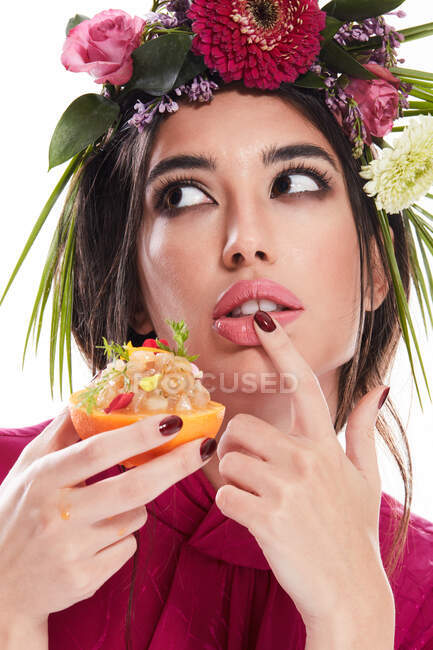 Young gorgeous woman with wreath of colorful flowers on head and finger on lip looking away while holding dish in orange zest isolated on white background — Stock Photo