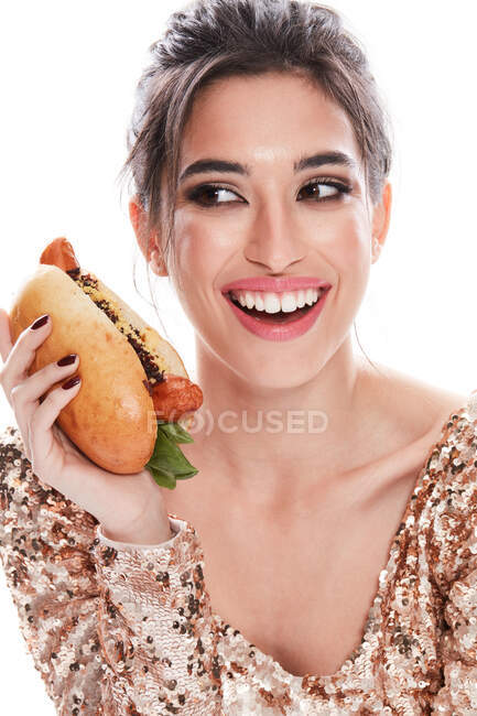 Glamorosa dama de pelo negro con un maquillaje elegante y la boca abierta mirando a la cámara mientras disfruta de hot dog aislado sobre fondo blanco - foto de stock