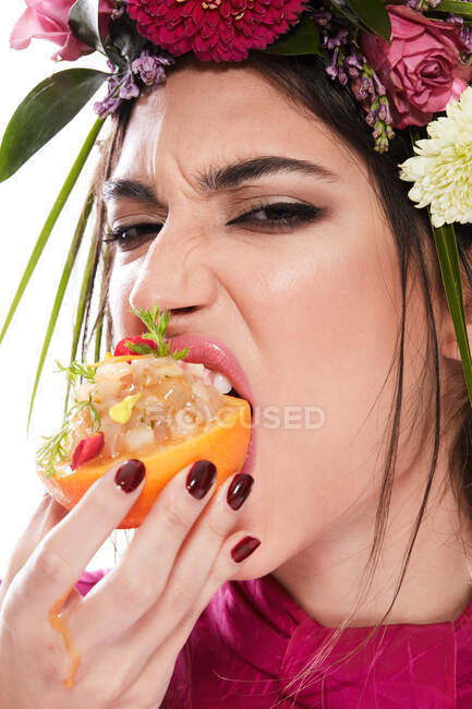 Giovane donna splendida con ghirlanda di fiori colorati sulla testa guardando la fotocamera mentre tiene piatto in scorza d'arancia isolato su sfondo bianco — Foto stock