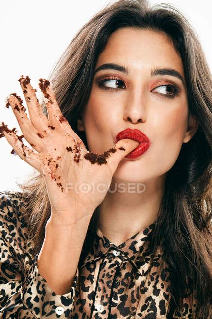 Attractive black haired female model with stylish makeup looking away while licking finger of hand smeared with cake cream isolated on white background — Stock Photo