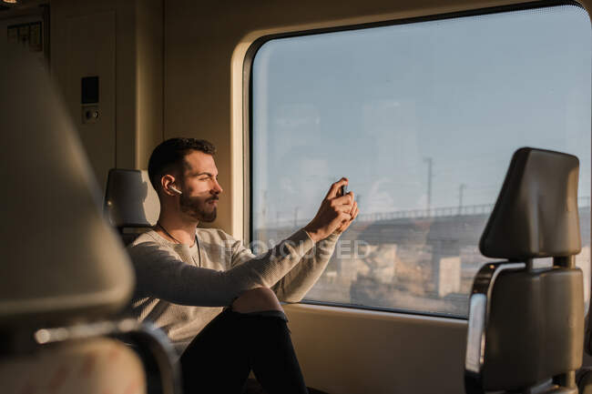 Concentrado jovem passageiro do sexo masculino fotografando no celular através da janela enquanto sentado no trem — Fotografia de Stock