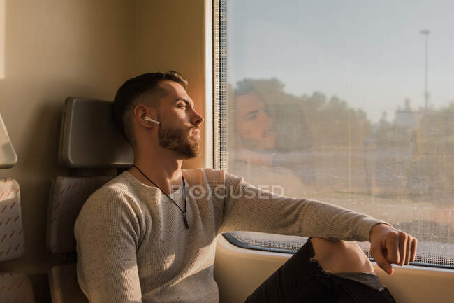 Pensive youthful bearded man with wireless earphones listening to music in train in sunny day — Stock Photo