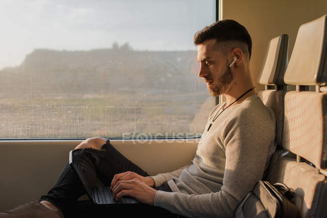 Side view of young man in wireless earphones typing on laptop during journey in subway in sunny day — Stock Photo