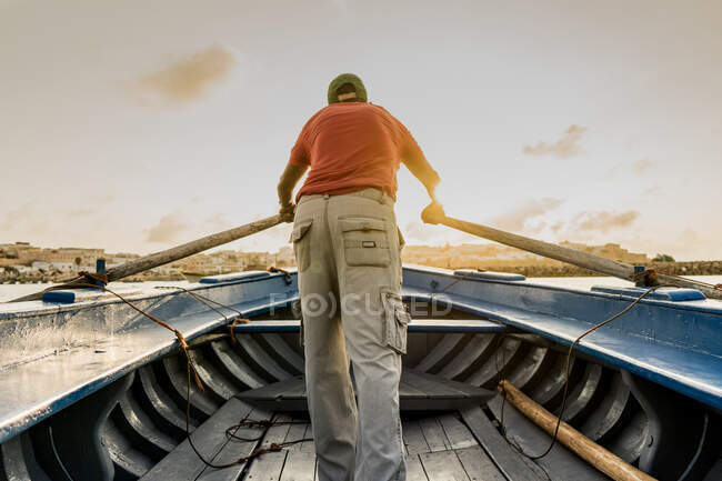 Vue arrière de l'homme sans visage en tenue décontractée debout dans un bateau en bois tenant de grandes rames dans les mains avec coucher de soleil doré sur le fond — Photo de stock