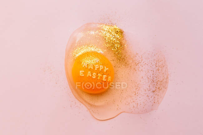 Draufsicht auf zerbrochenes rohes Ei mit Text Frohe Ostern auf Eigelb bedeckt mit goldenen Funkeln auf rosa Hintergrund — Stockfoto