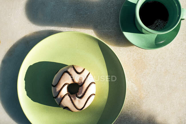 Сверху чашка вкусного кофе на блюдце рядом с вкусным пончиком с шоколадной глазурью на зеленой круглой тарелке на сером столе — стоковое фото
