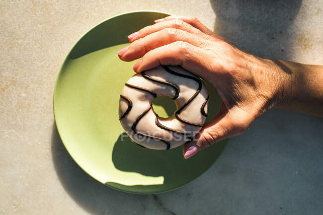 De arriba cosecha hembra sosteniendo rosquilla deliciosa con glaseado por encima del plato de cerámica verde mientras desayuna en la terraza de la cafetería en el día soleado - foto de stock
