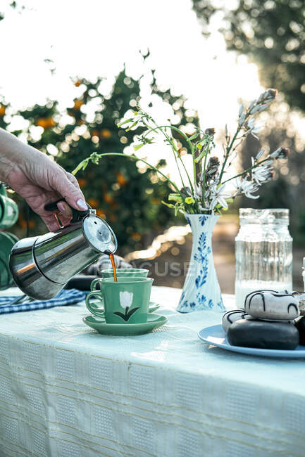 Cosecha femenina verter café de metal géiser cafetera a taza verde mientras se prepara el desayuno de pie en la mesa con ramo de flores silvestres en jarrón y rosquillas frescas con esmalte en el plato en la naturaleza - foto de stock