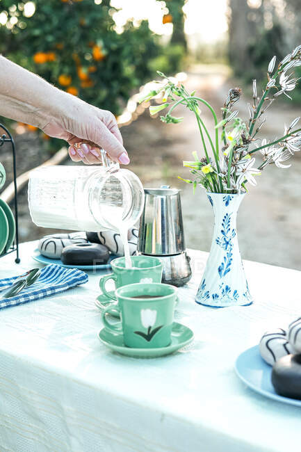 Mujer vertiendo leche fresca de jarra a taza de cerámica mientras hace un picnic en el jardín - foto de stock