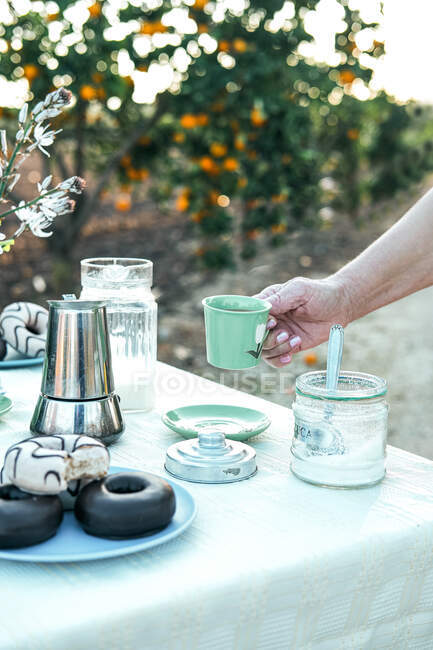 Mujer con taza de cerámica verde de bebida sabrosa desayunando en la mesa con rosquillas frescas en el plato junto a varios vajilla en la naturaleza - foto de stock