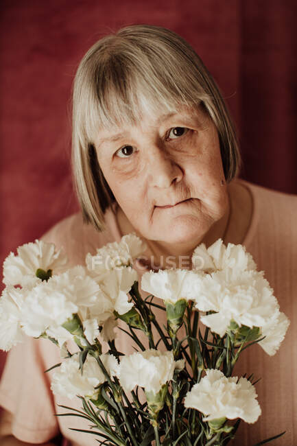 De cima pensativo velha fêmea com cabelos grisalhos olhando para câmera e segurando buquê de cravo branco em casa — Fotografia de Stock