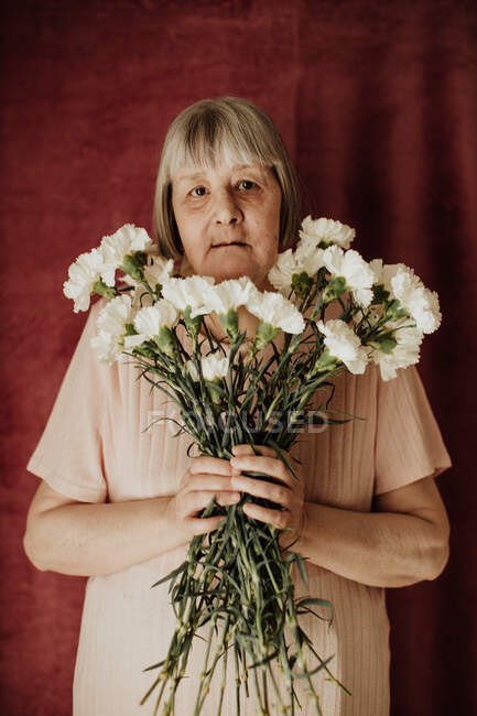 Зверху вдумлива стара жінка з сірим волоссям дивиться на камеру і тримає букет білої гвоздики вдома — стокове фото