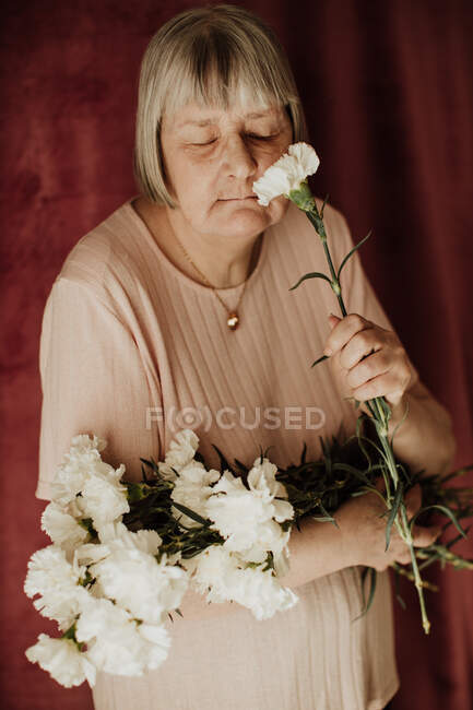 Desde arriba pensativa vieja hembra con el pelo gris con los ojos cerrados sosteniendo ramo de clavel blanco en casa - foto de stock