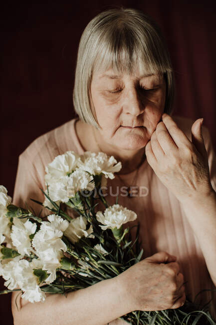 From above thoughtful old female with gray hair with closed eyes holding bouquet of white carnation at home — Stock Photo